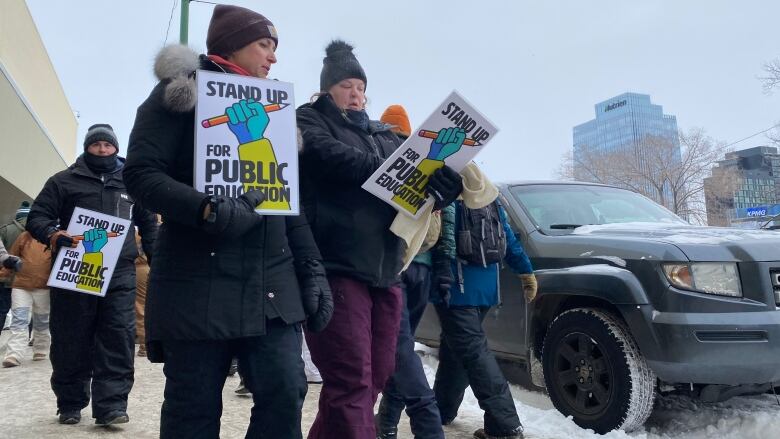 teachers dressed in winter clothes walk along sidewalk with posters that say 