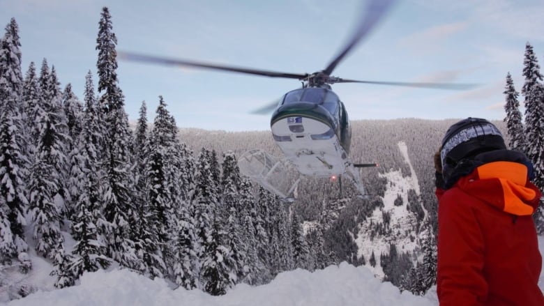 A helicopter touches down on a wintry mountainside. A skier can be seen from behind.