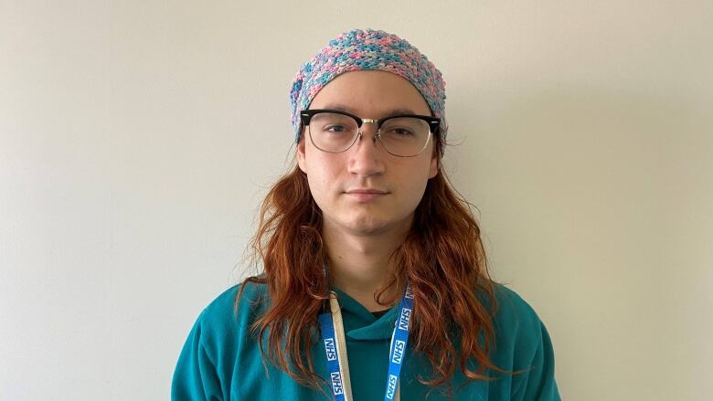 a person with long red hair wearing a teal hoodie, knitted hat and glasses.