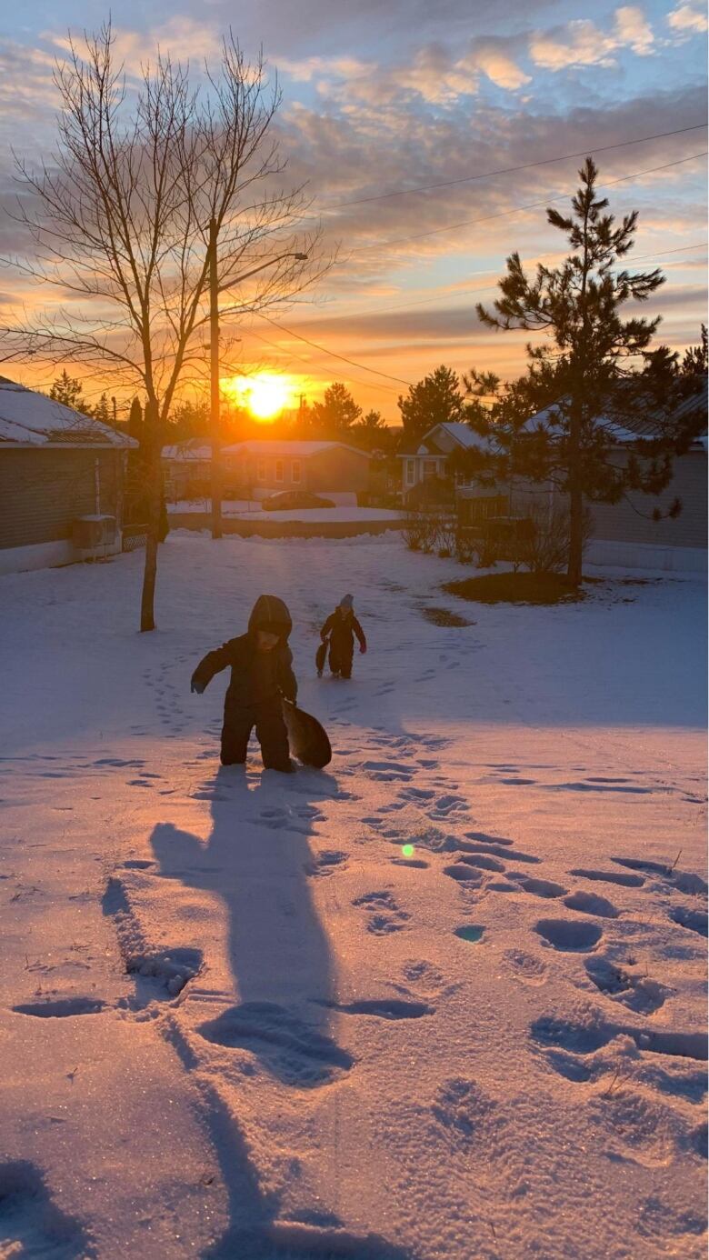 Two small children dressed in snowsuits run through a snow-laden yard while the sun sets in the distance.