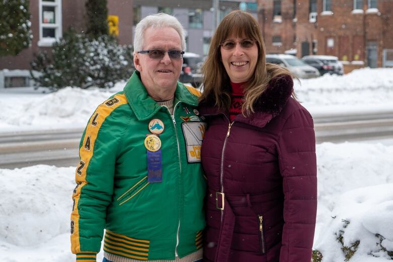 A man with white hair, wearing a green and yellow 'North End Blazers' jacket stands beside a woman with brown hair wearing a dark pink parka on a snowy sidewalk. They both smile at the camera. 