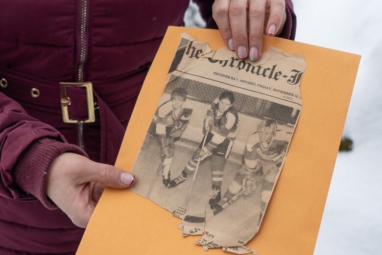 A close-up of an old newspaper photo that's seen some wear and tear. It shows a young female hockey player flanked by two male teammates. 