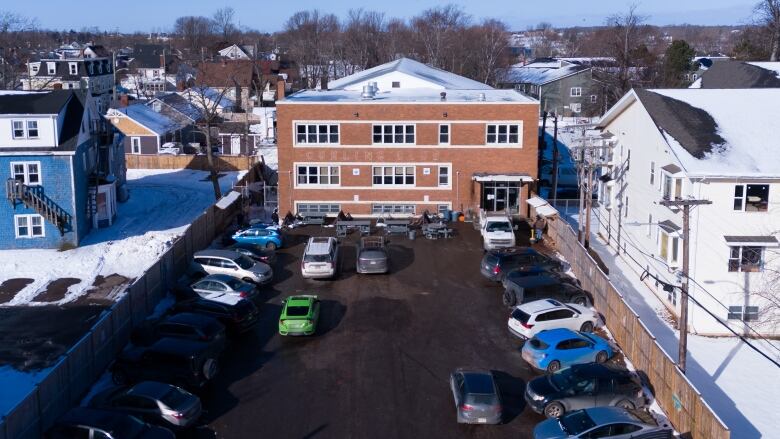 A three-story brick building is shown from the air, with cars filling its parking lot and picnic tables near the front door. 