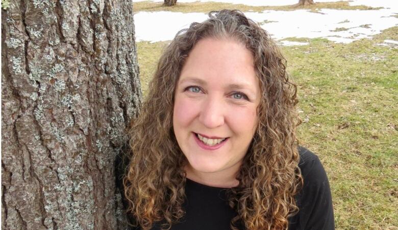 Woman with curly brown hair smiles next to a tree trunk