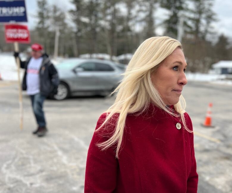 Woman in red winter coat, with man in background holding Trump signs.