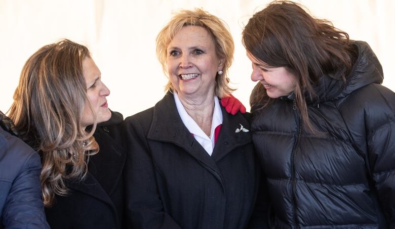 three women standing in a row, wearing winter jackets. the two at each end are looking at each other.