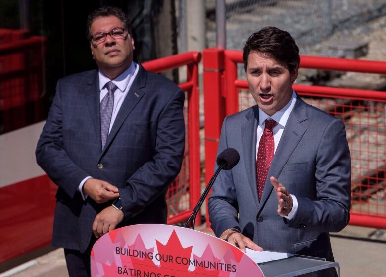 A suited man speaks behind a Canada flag lectern, while another man looks on behind him. 