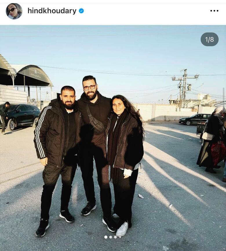 A man with a dark beard and glasses, wearing a black hoodie and pants, stands on a road between a man on his left and a woman on his right.