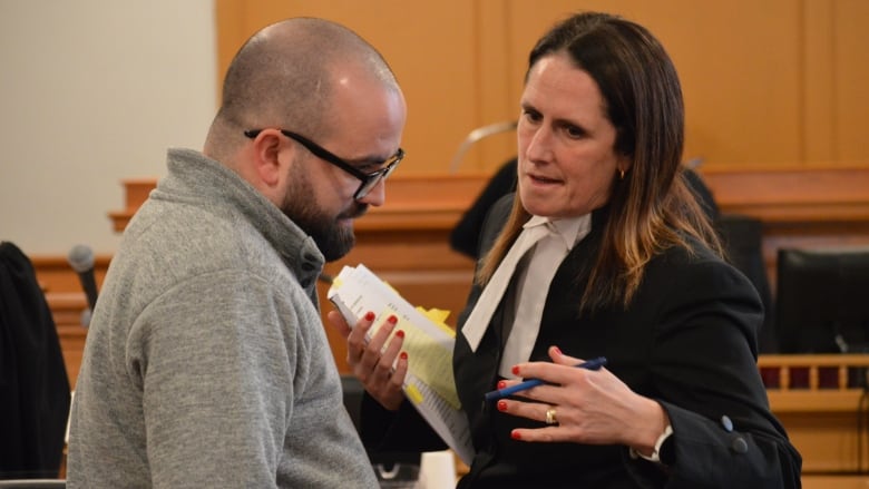 A woman in court attire speaks privately to a bald man with a beard and glasses in a courtroom