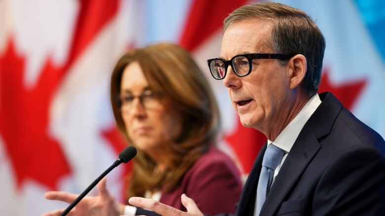  A man with glasses, wearing a suit, speaking into a microphone