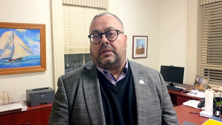 A white man with short grey hair, stubble and glasses wears a grey blazer while standing in an office