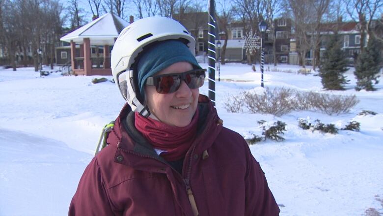 Woman wearing a helmet and sunglasses smiling wearing burgundy winter coat, standing outdoors on an ice rink.