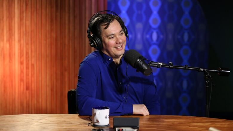 A man wearing a blue button-up shirt, the playwright David Yee, sitting at a wood table in front of a studio microphone, wearing headphones.