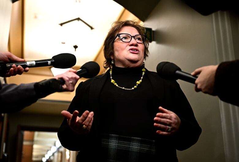 A woman wearing a black turtle neck and gold necklace speaks as reporters hold microphones.