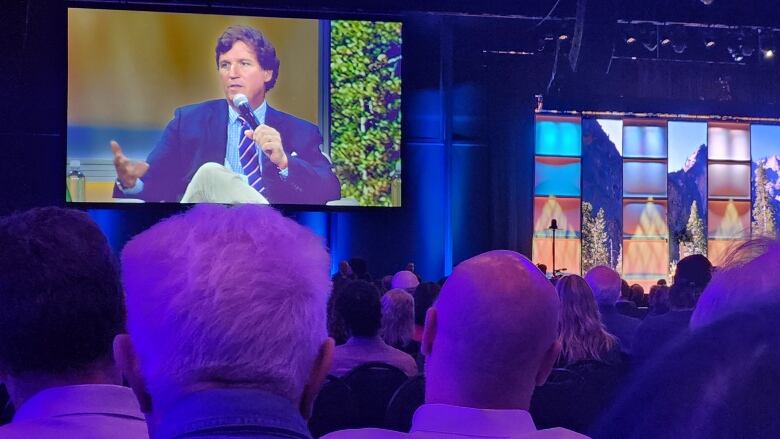 People face a stage and large video monitors of a man talking.