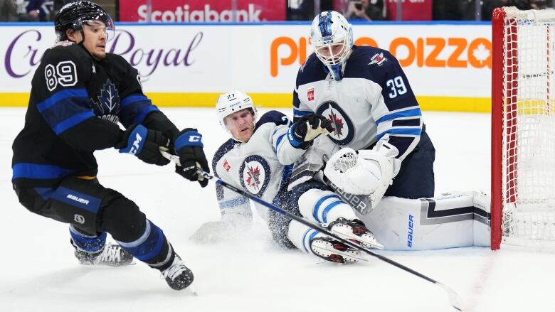 A hockey player slides across the ice in front of a goalie as a player from the opposing team reaches his stick toward the puck.