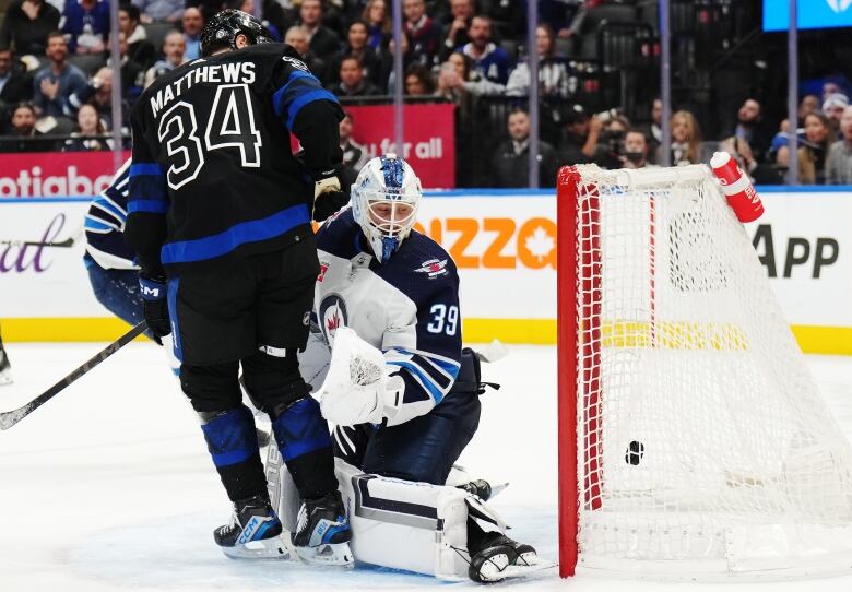 A hockey puck hits the back of a net as a goalie looks over his shoulder and another player from the opposing team stands in front of him.