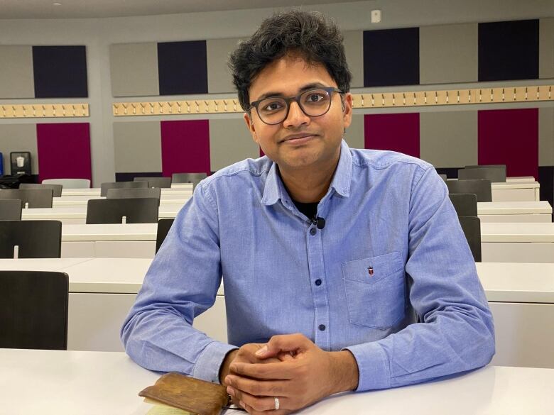 A young man sits at a desk in a lecture room and smiles at the camera.