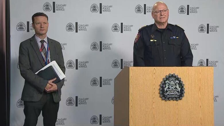 two men stand in front of a podium. one man is wearing a suit, the other in a police uniform.