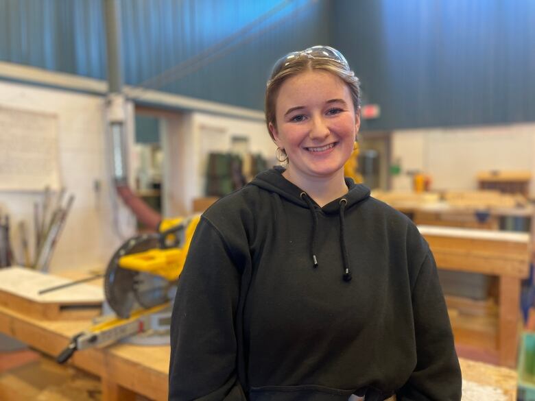 A girl poses smiling for a photo. She is in a black sweater and standing in a carpentry classroom with wooden tables behind her. 