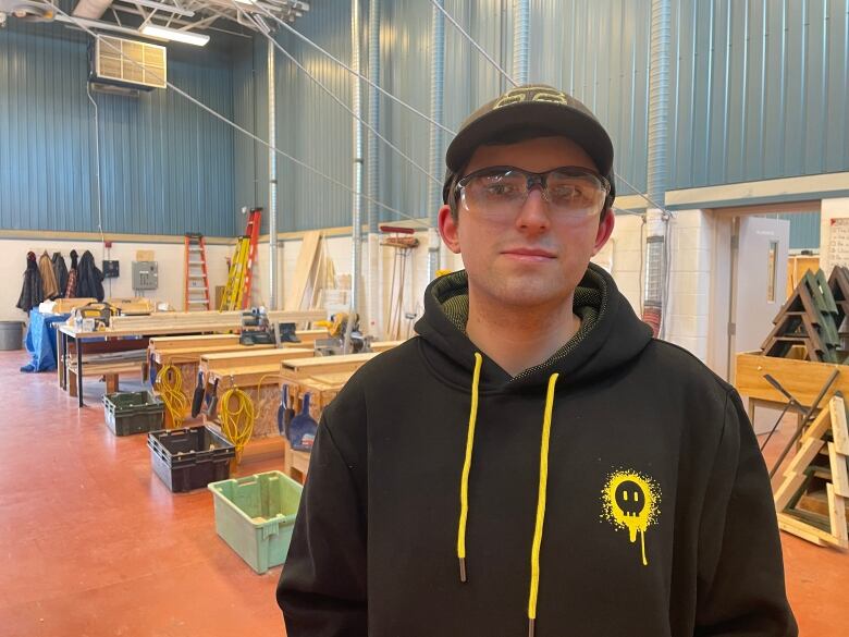 A Zach Lapointe poses for a photo. He is in a carpentry classroom. Behind him are wooden work tables. He is wearing a grey ball cap, safety glasses and a black hoodie.