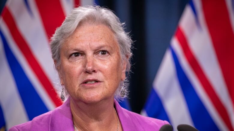 A white woman with white hair and wearing a pink blazer stands in front of two B.C. flags at a podium.