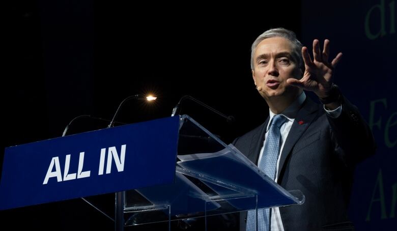 A man at a podium raises one hand against a black background.