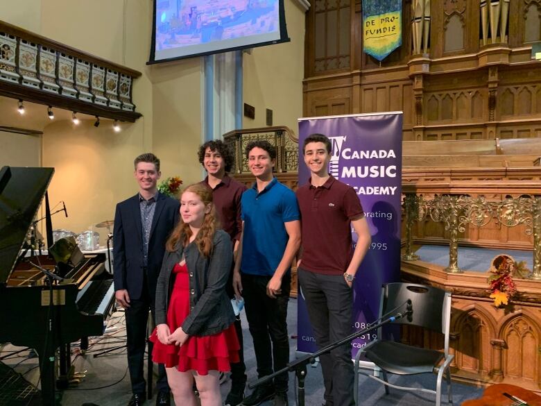 A group of teenagers stand in a church with a piano seen in the background. 