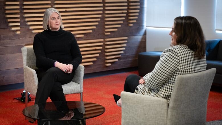 An older woman with gray hair and a black sweater sits on a gray seat. To the right of the frame is a woman with brown hair, wearing a checkered blazer and sitting on another gray chair.