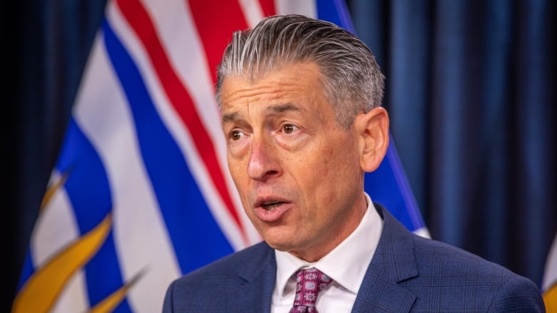 A white man wearing a purple tie stands in front of B.C. flags.