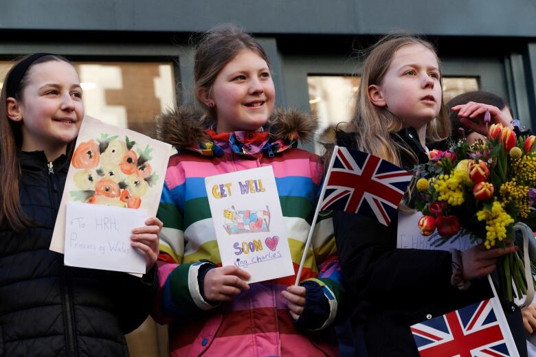 Three children standing beside one another hold greeting cards, flowers and flags.