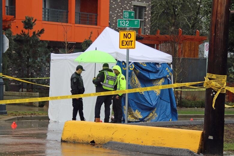 Three officers stand under an umbrella with a white tent behind them. 