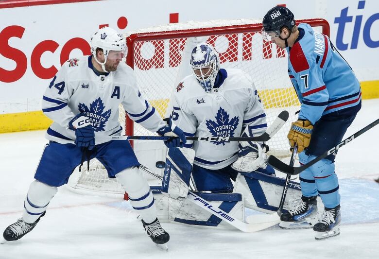 Two players jockey for position in front of a goaltender as the puck enters the fray.