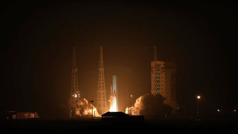 A night view of a rocket blasting off from a launch pad surrounded by towers.