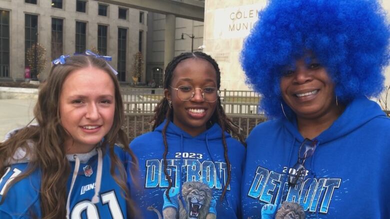 Three women wearing blue sports memorabilia with the woman on the far right wearing a wig of a blue afro