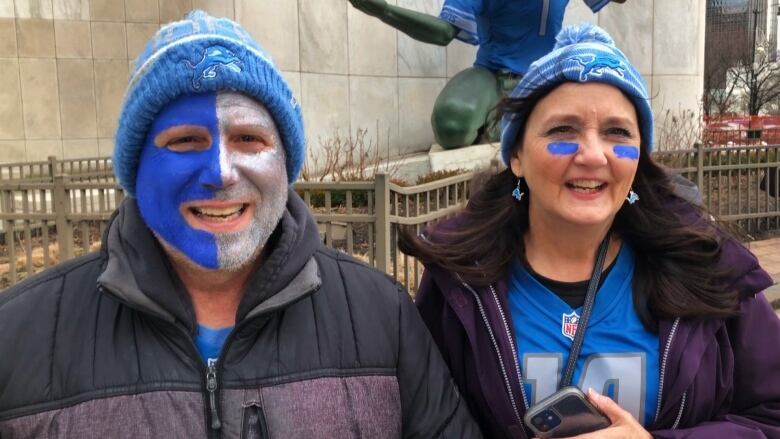 A couple wearing blue sports jerseys under black jackets and blue and white face paint