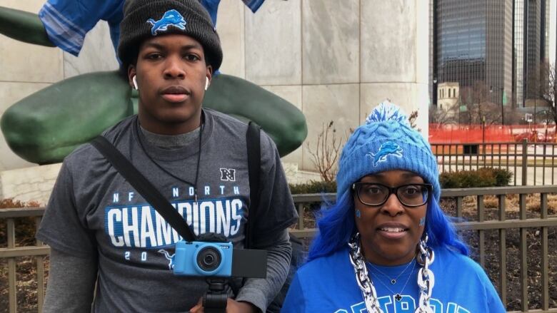 A tall boy wearing two grey shirts and a black hat standing beside his mother wearing a blue hat and a blue shirt