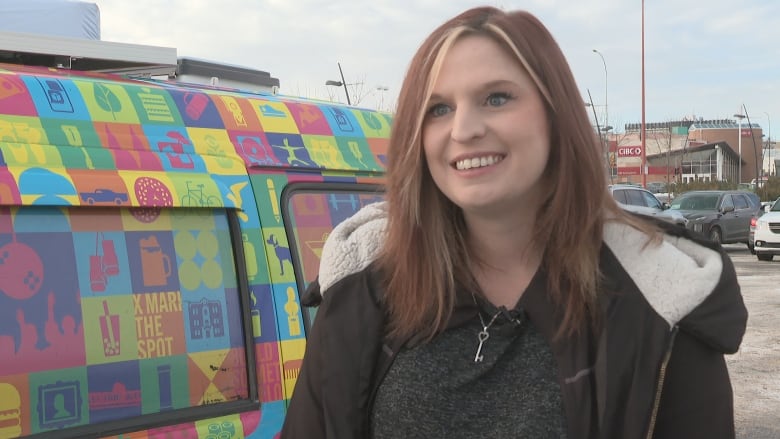 A woman in a black jacket and with brown hair is seen smiling as she stands in front of a colorful van.