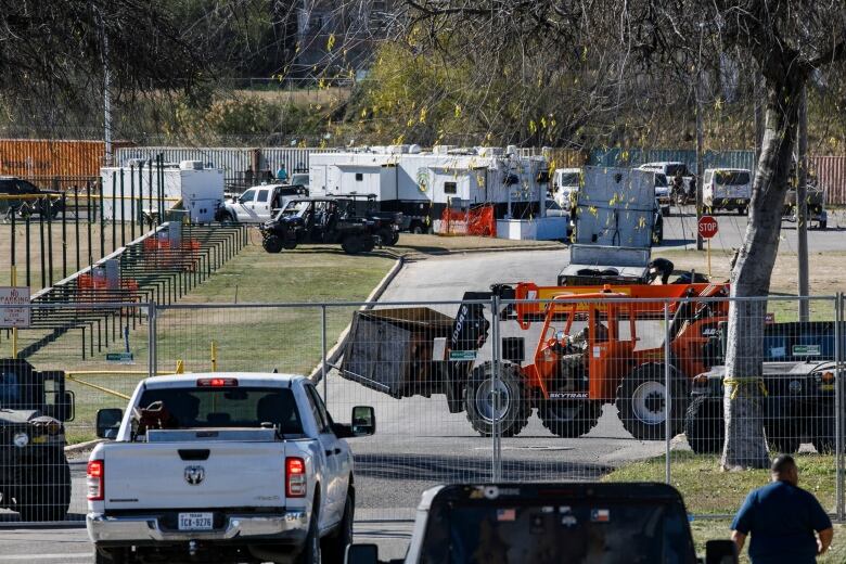 A white pick-up truck parked in front of a metal fence with an orange forklift parked behind and several white trailers in the background.