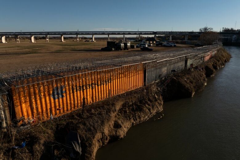 A line of large shipping containers, with barbed wire on top, stand along a river bank. 