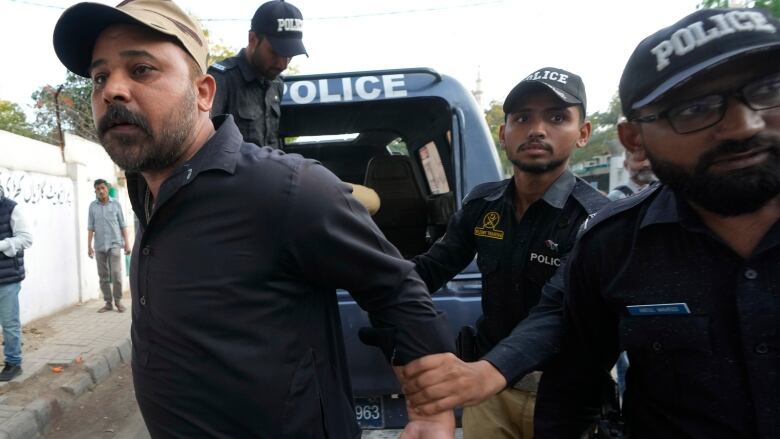 A bearded man in a baseball cap is shown getting his wrists placed behind me in apparent arrest by a police officer.