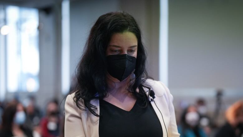 A woman with long black hair wearing a cream coloured blazer and a black face mask holds a stack of papers as she walks toward a podium.