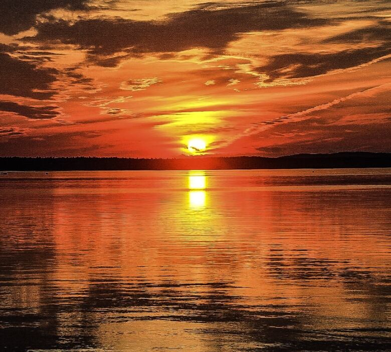 A burning orange sky reflects its colours across a lake. Trees silhouetted black line the horizon. 