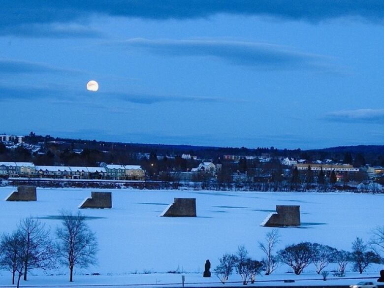 A full moon sits halfway-up a deep blue sky. Underneath are houses nestled along a snowy river.