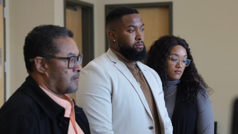 Three people stand with solemn facial expressions.