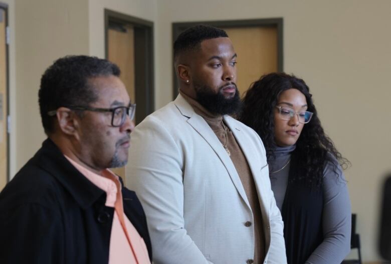 Three people stand with solemn facial expressions.