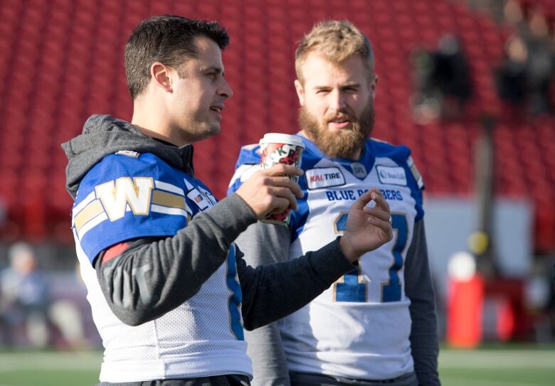 Two football players in white and blue uniforms stand beside one another in conversation, without helmets.