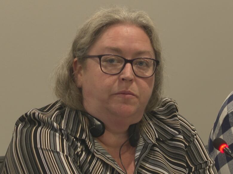 A woman with light brown shoulder length hair, parted in the middle and wearing dark rimmed glasses and a striped black, gray and brown blouse, sits at a table in front of a microphone.