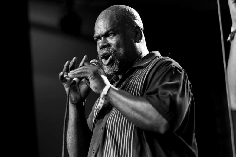 A bald man with a goatee is singing into a microphone and wearing a black collared shirt. The photo is black and white. 