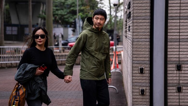 A man in a green hooded jacket is seen walking, holding hands with a woman wearing sunglasses.